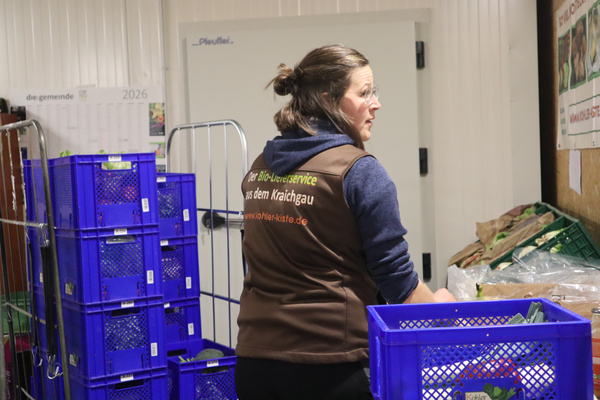 KI generiert: Eine Frau sortiert Kisten in einem Lager. Text auf Weste: "Der Bio-Lieferservice aus dem Kraichgau www.bioher-kiste.de".