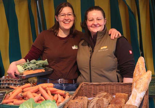 KI generiert: Zwei Frauen stehen hinter einem Markttisch mit Gemüse und Brot und lächeln.