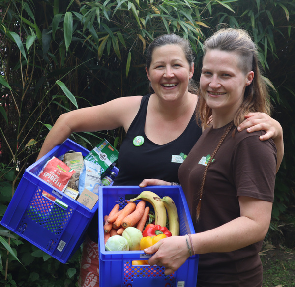 KI generiert: Zwei Frauen halten blaue Kisten mit Obst, Gemüse und Lebensmitteln in einem Garten.