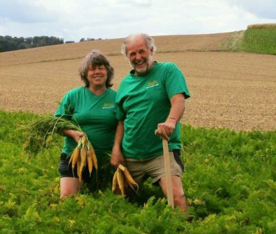 KI generiert: Zwei Personen stehen auf einem Feld mit Karotten in der Hand, lächelnd in die Kamera.