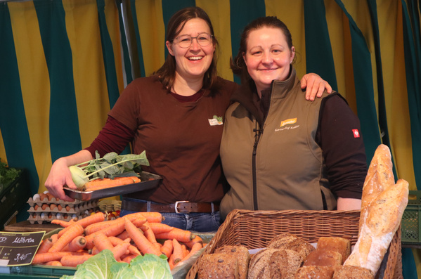 KI generiert: Zwei Frauen stehen lächelnd hinter Gemüse und Brotständen. Text: "Möhren 3,45€".
