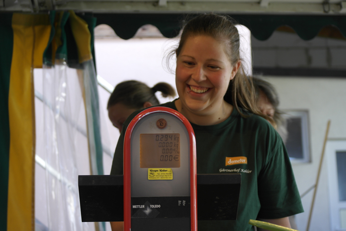 KI generiert: Eine lächelnde Frau steht hinter einer Waage. Text: "demeter, Gärtnerhof Keller".