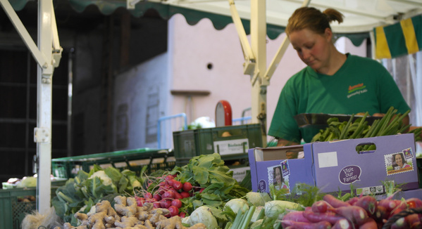 KI generiert: Marktstand mit frischem Gemüse und einer Verkäuferin im grünen T-Shirt. Text: "Visit Your Farmers".