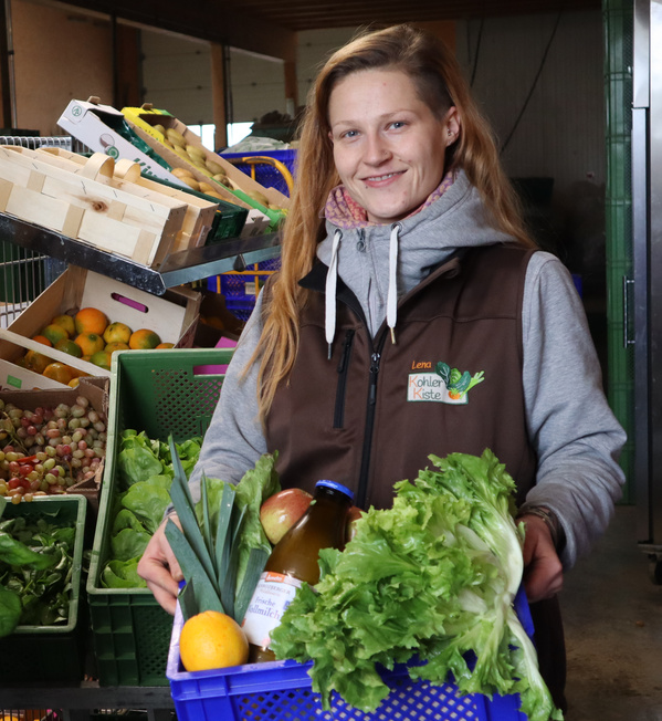 KI generiert: Eine Person hält eine Kiste mit frischem Gemüse und Obst, umgeben von weiteren Kisten.