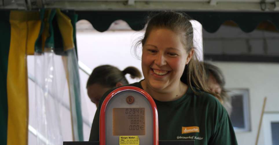 KI generiert: Eine lächelnde Frau steht hinter einer Waage mit Display. Text auf Tafel: "Demeter Gärtnerhof Keller".