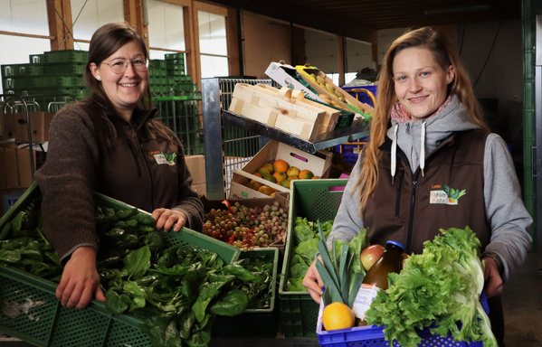 KI generiert: Zwei Personen halten Kisten mit frischem Gemüse und Obst in einem Innenraum.