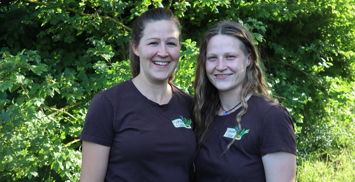 KI generiert: Zwei Personen in braunen T-Shirts stehen vor einem grünen Busch und lächeln in die Kamera.