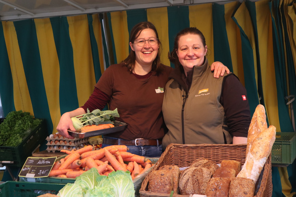KI generiert: Zwei Frauen an einem Marktstand mit Gemüse, Brot und Schildern. Text: "Möhren, 3,45€".