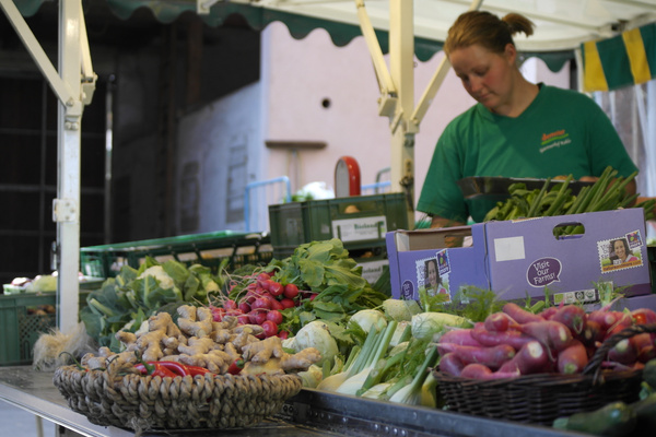 KI generiert: Ein Marktstand mit frischem Gemüse und einer Verkäuferin, die das Angebot ordnet.